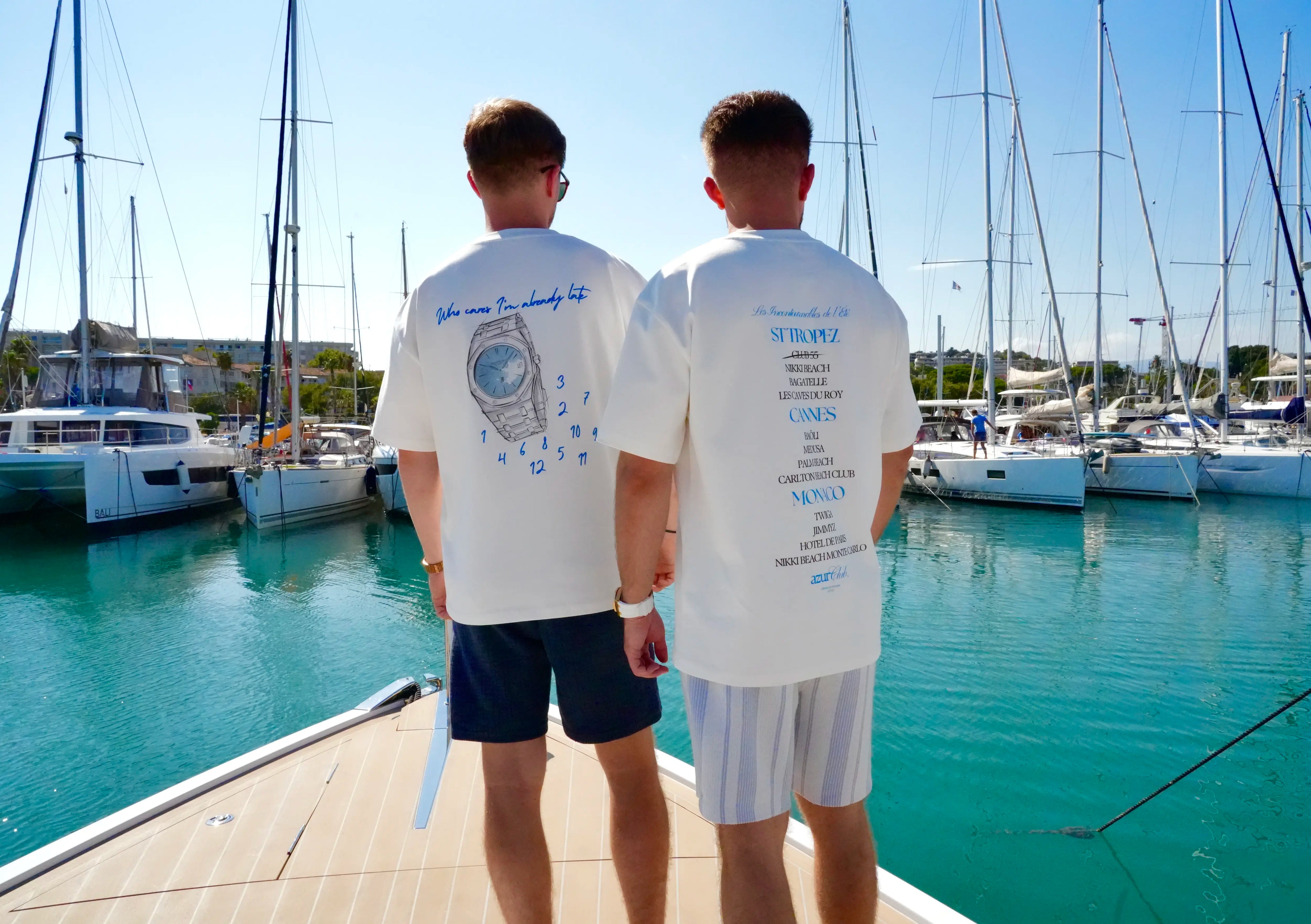 Two people wearing matching white t-shirts with text and graphics, standing on a dock with boats in the background.